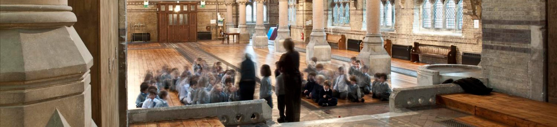 A group of school children sat on the floor of an empty church building which has arches decorated with gold, a runway of tiles in a pattern, carved stonework and a circular stained glass window. The people in the image are blurred by a long exposure and are unidentifiable.