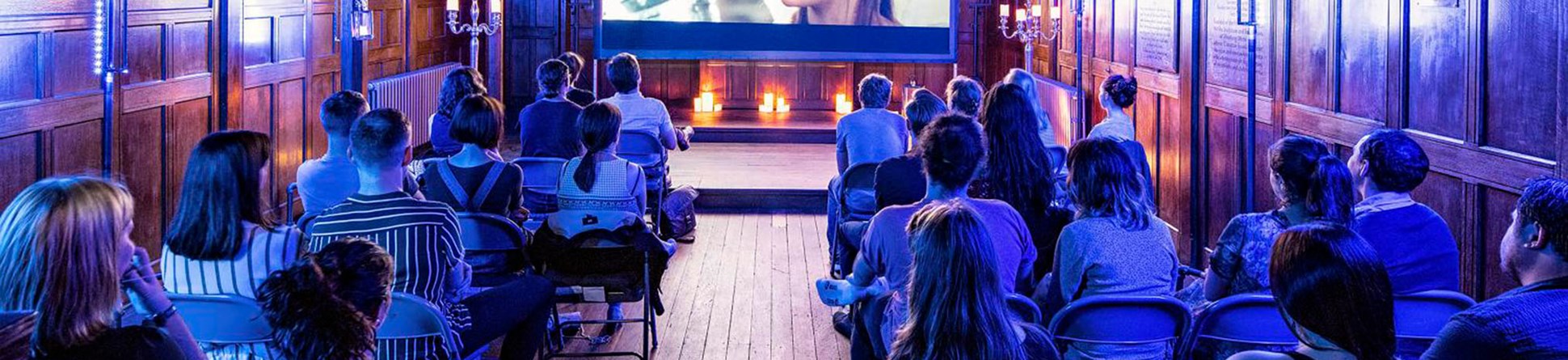 An audience watching a film in a wood-panelled hall with blue lighting and a large screen displaying a movie scene.