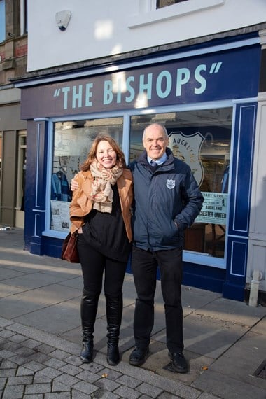 A woman and man standing on the pavement in front of a shop, the shop sign reads "The Bishops".
