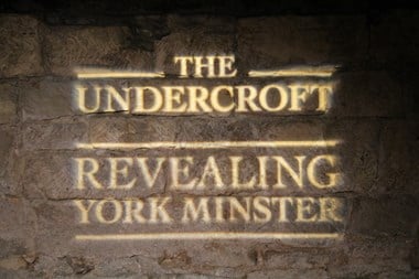 Image of a sign projected onto the stone wall of the Undercroft Gallery entrance. The sign reads: The Undercroft, Revealing York Minster