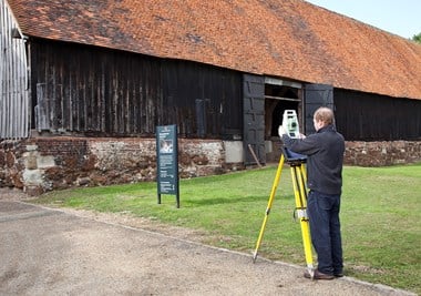 Colour photograph of a man setting up a total station theodolite on a tripod in front of an old barn