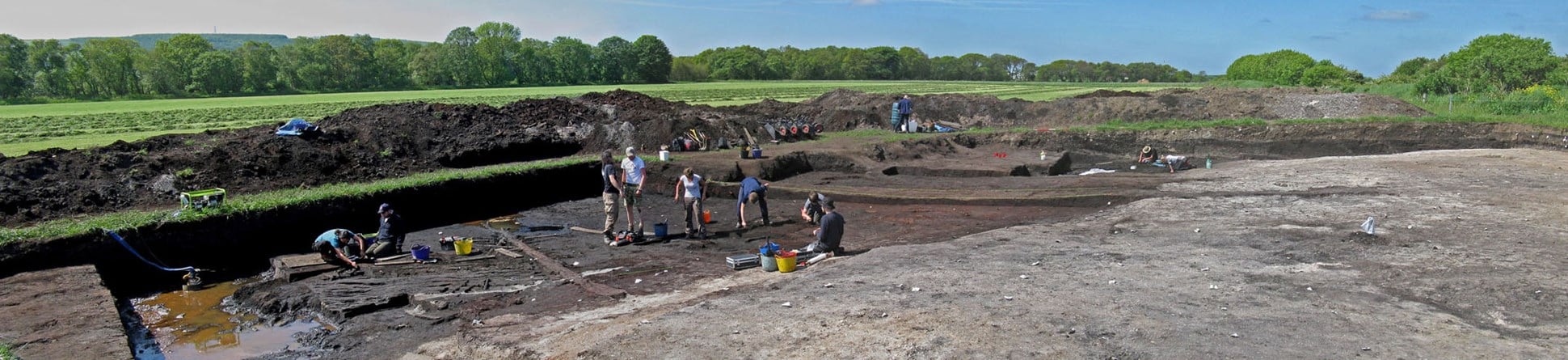 Colour photograph showing lots of archaeologists digging in a trench with spoilheaps behind; some timbers are visible left