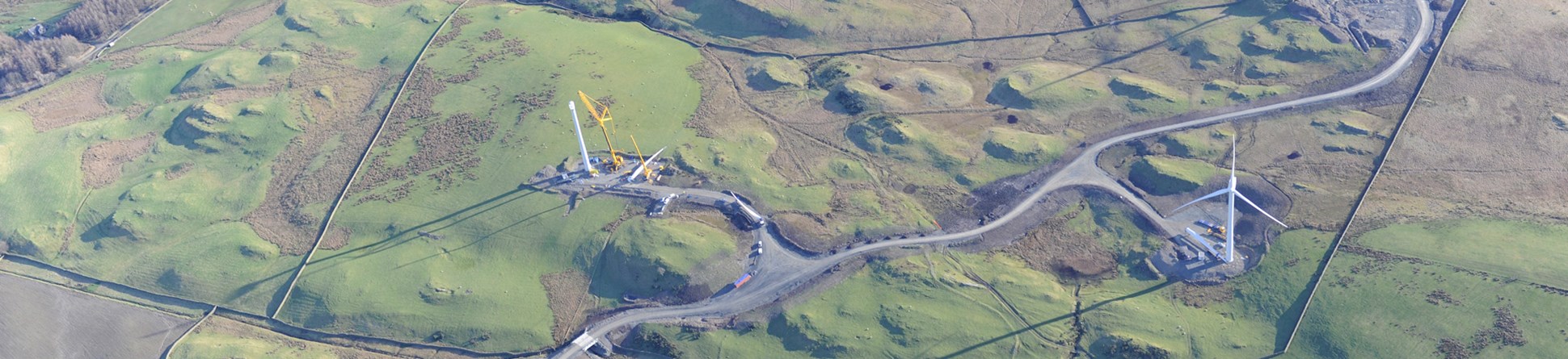 Colour aerial photograph showing one complete wind turbine and one under construction with the cranes and construction road