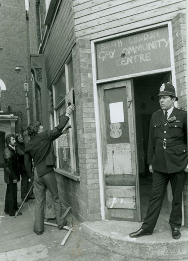 Black and white photo of a policeman standing in the doorway of the South London Gay Community Centre while three men look at notices in the window to the left of the door.