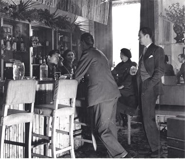 The photograph shows Ian Board, Colony Room bartender and later proprietor (seated behind bar), Carmel Stuart, Muriel Belcher's lover (seated behind bar), Nina Hamnett, artist and bohemian (seated at bar)