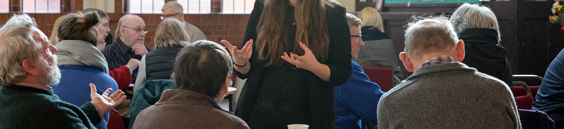 A person with long brown hair stands and gestures while speaking to seated elderly people in a community hall with tea and coffee cups.