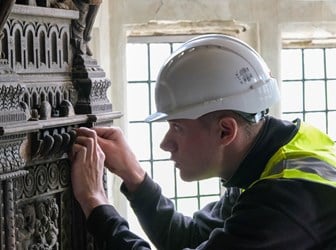 A photograph of a man in a white helmet and hi-vis jacket closely inspecting a decorative wooden object in front of a window.