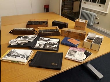 A colour photograph of three tables pushed together. On the tables are open and closed photograph albums, wood, and card boxes of photographic slides, books, and magazines.