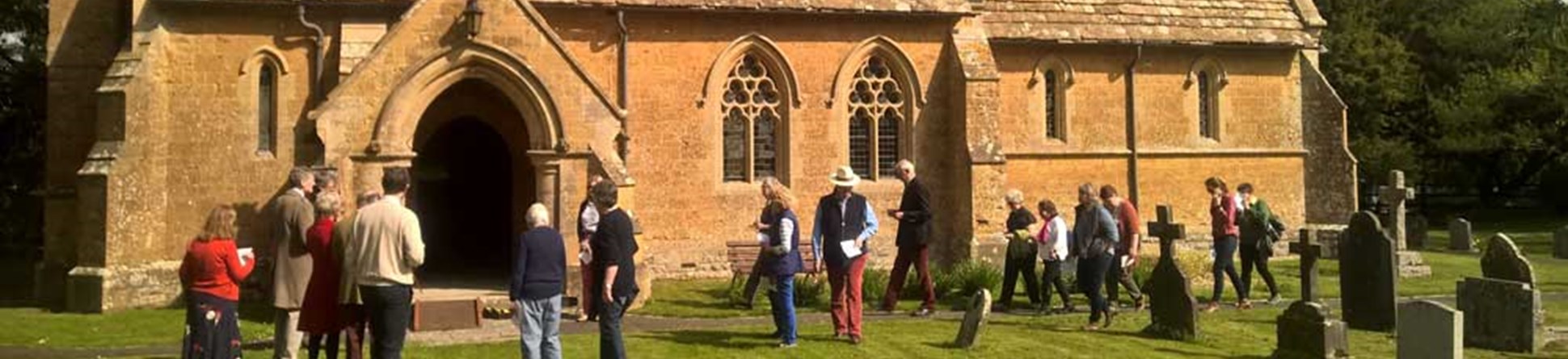 A photograph of people gathering outside a small, honey-coloured church set in a well-kept graveyard.