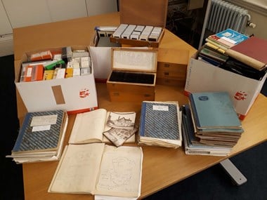 A colour photograph of three tables pushed together. On the tables are cardboard boxes containing boxes of photographic slides, open and closed notebooks, and wooden boxes containing different sized photographic slides.