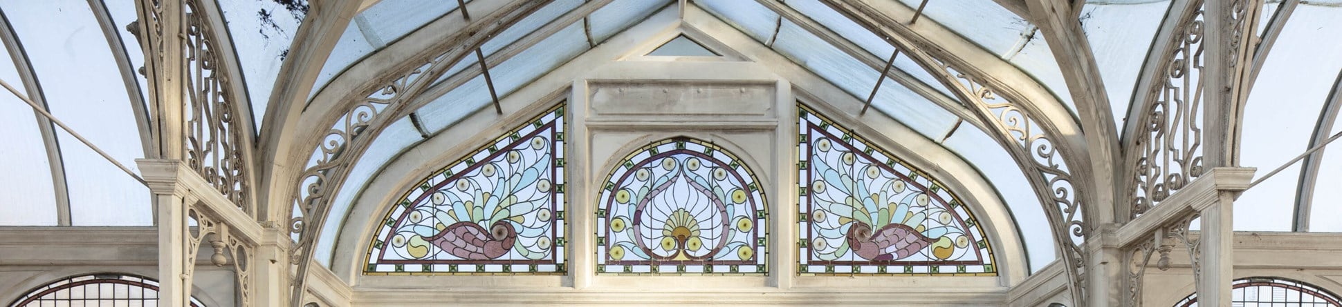 A white framed conservatory with decorative scrolled ironwork and, above plain glass windows, three beautifully decorated panels showing birds and a central peacock feather design.