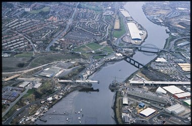 A colour oblique aerial photograph showing a river with road and rail bridges crossing it. In the foreground, a third bridge is under construction. Roads and industrial buildings line the riverbanks. In the top left quarter of the photograph are streets of houses.