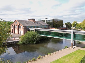 A glass-enclosed walkway crossed a canal to reach a red brick building, with a modern glass building behind.