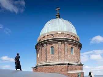 A figure in a priest's cassock stands on a curved roof, looking at the brickwork and copper roof of a domed church.