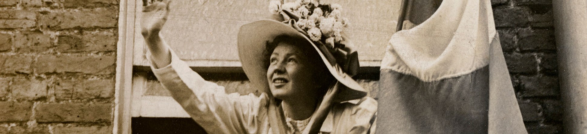 Christabel Pankhurst waving out of a window, with a flag in her left hand, wearing large straw hat with flowers on top.