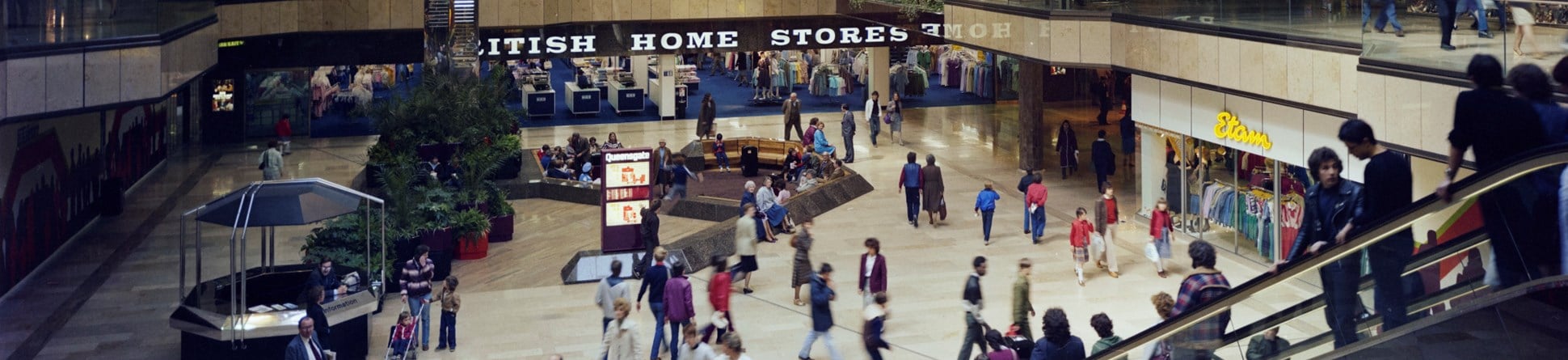 Colour photograph showing shoppers inside Queensgate shopping centre. The two levels of British Home Stores are in the background. On the right, shoppers are riding down an escalator into a large central square.