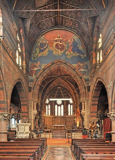 View of the altar with a painting of Jesus and his apostles directly above it. The walls are composed of red and black bricks.