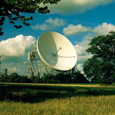 A gigantic dish looms over trees and agricultural fields.