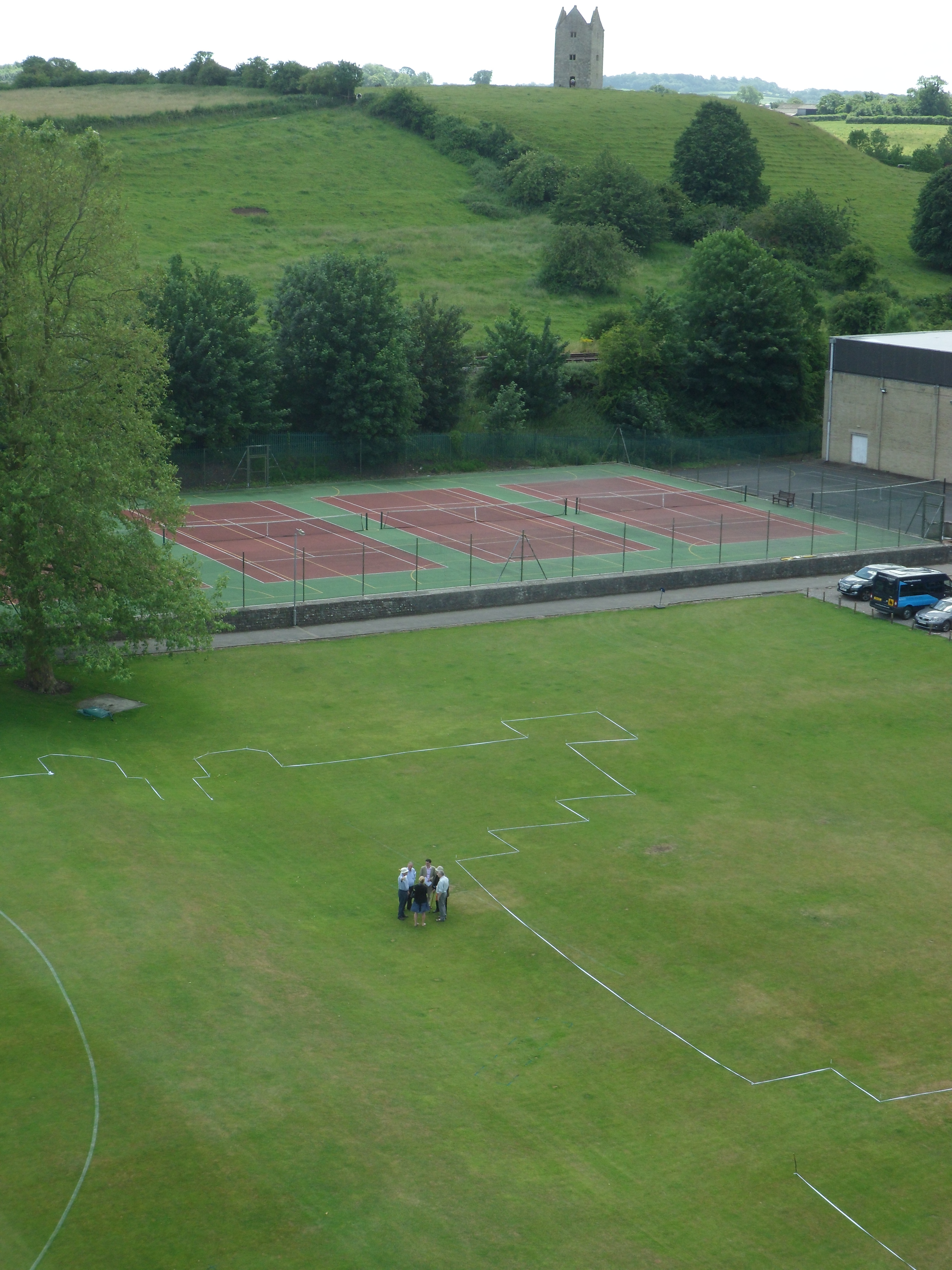 Small goup of people within a footprint of the mansion marked with white line on grass seen in front of tennis courts.