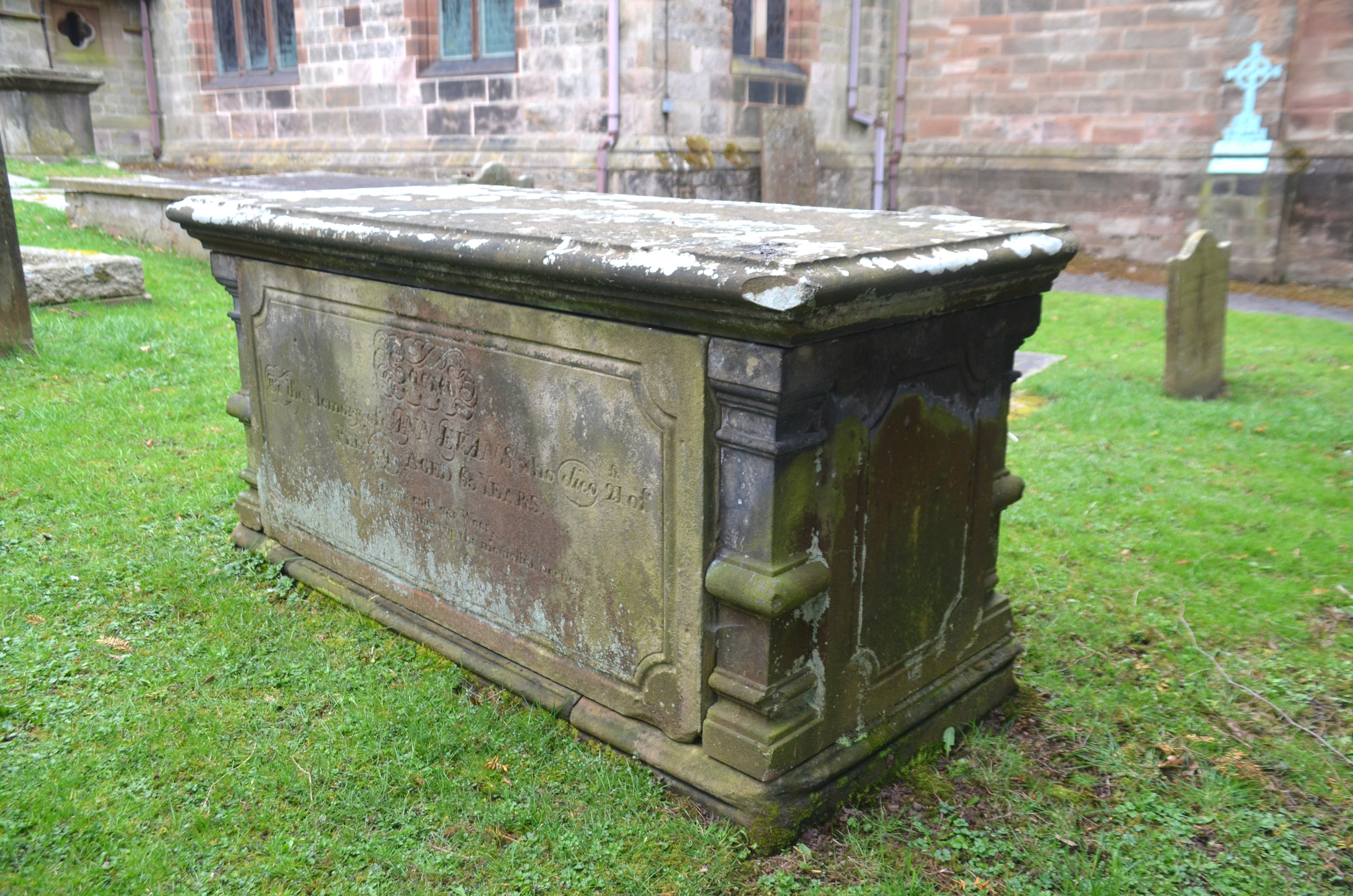 View showing inscribed south side panel and east end panel. Green grass with south aisle of church behind