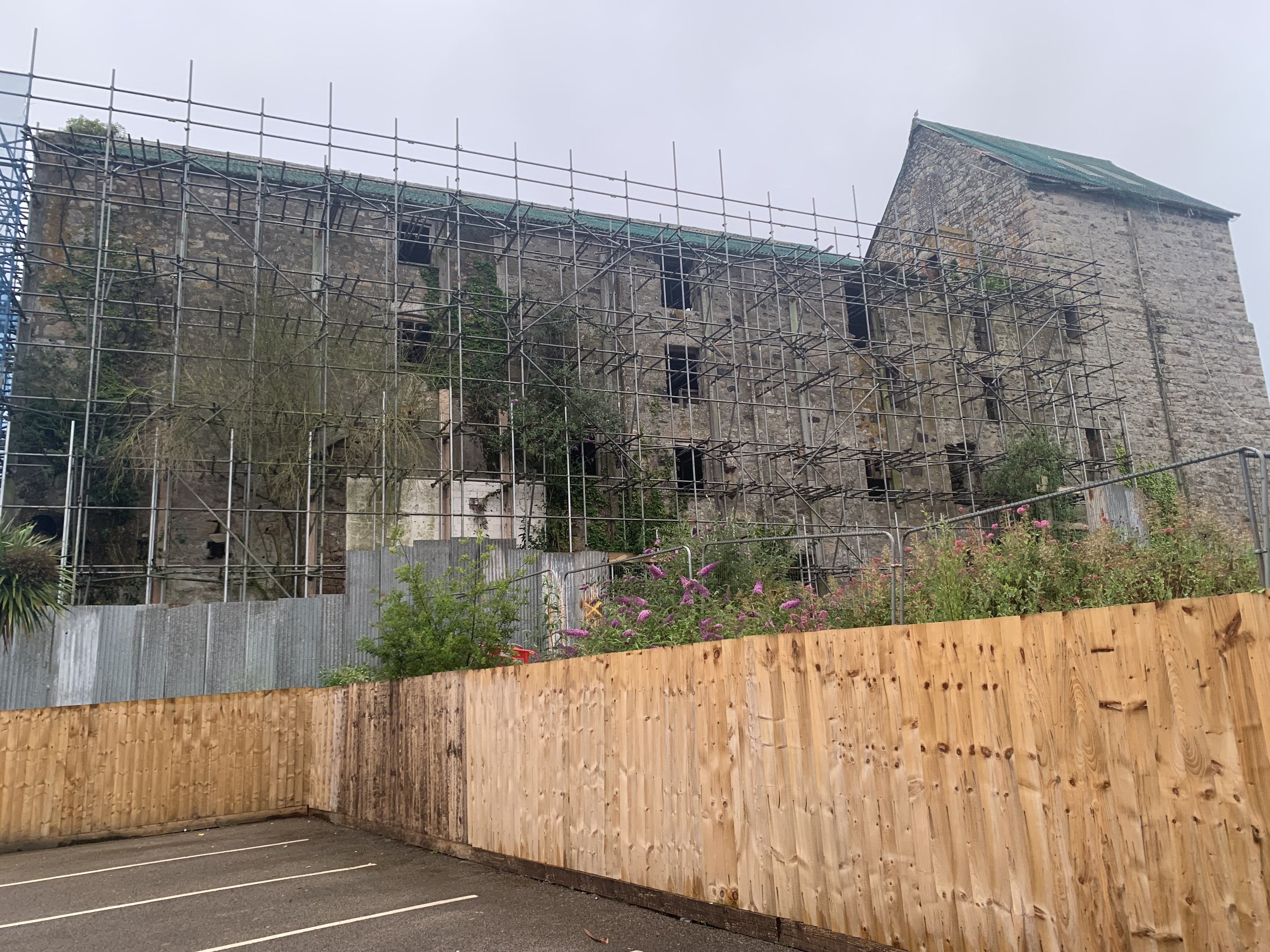 Old stone building, with all elevations covered in scaffold. Roof has grey sheet material, with green netting over it