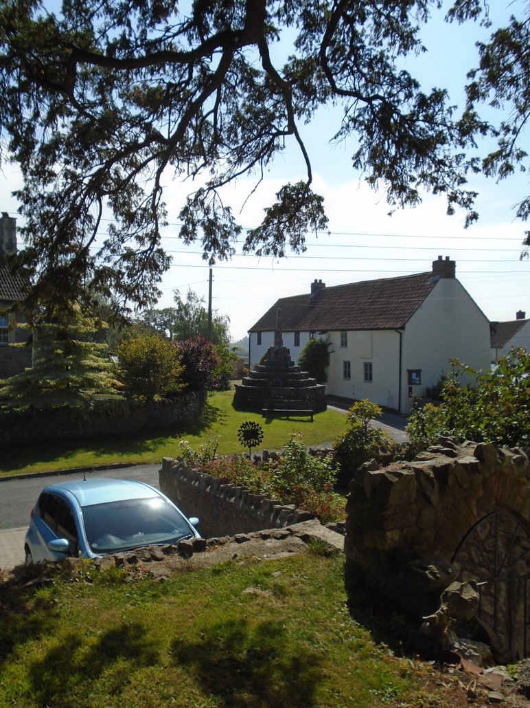 The east front of Banksea Cottages seen  across the road from the churchyard, with the massive cross base in front.