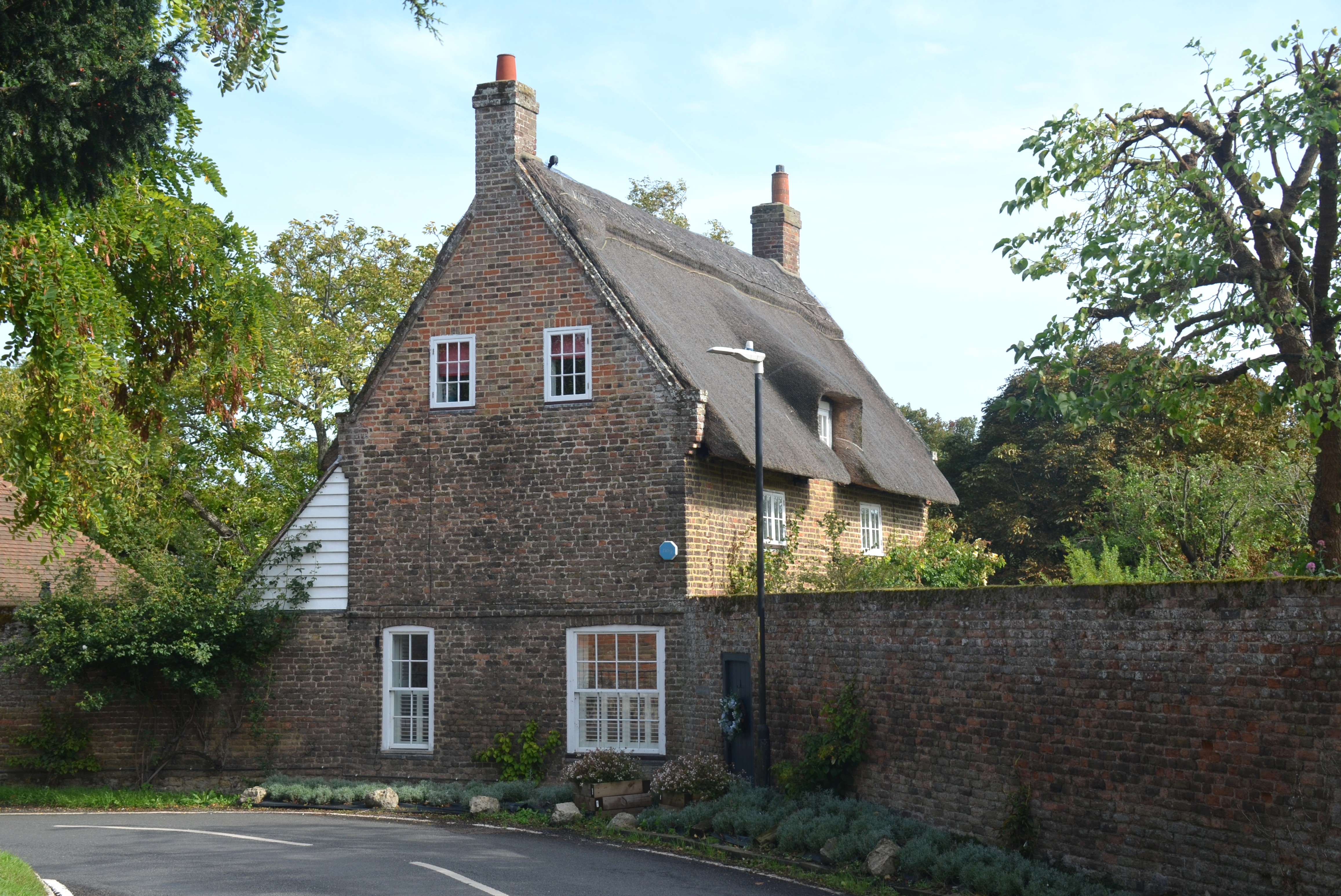 Thatched roof. Wall and windows of ground floor at street level. Trees to left.