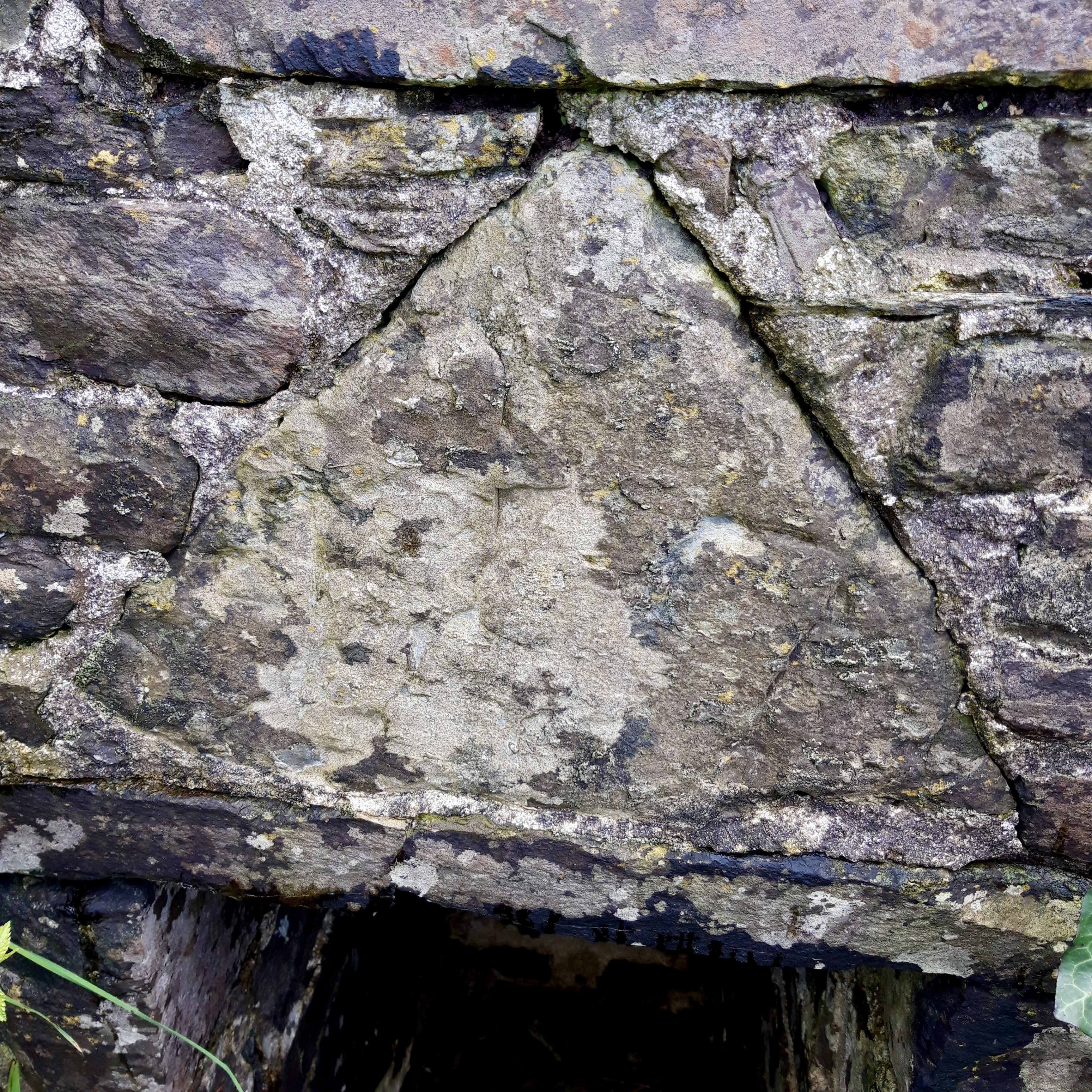 Photograph shows a large inscribed triangular stone incorporated into the structure of the wellhouse. 