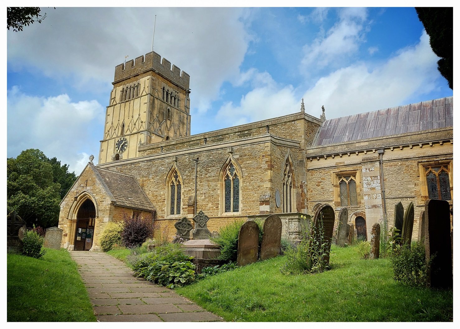 A colour photograph of a very old church in yellow stone, framed by trees and with blue sky beyond.