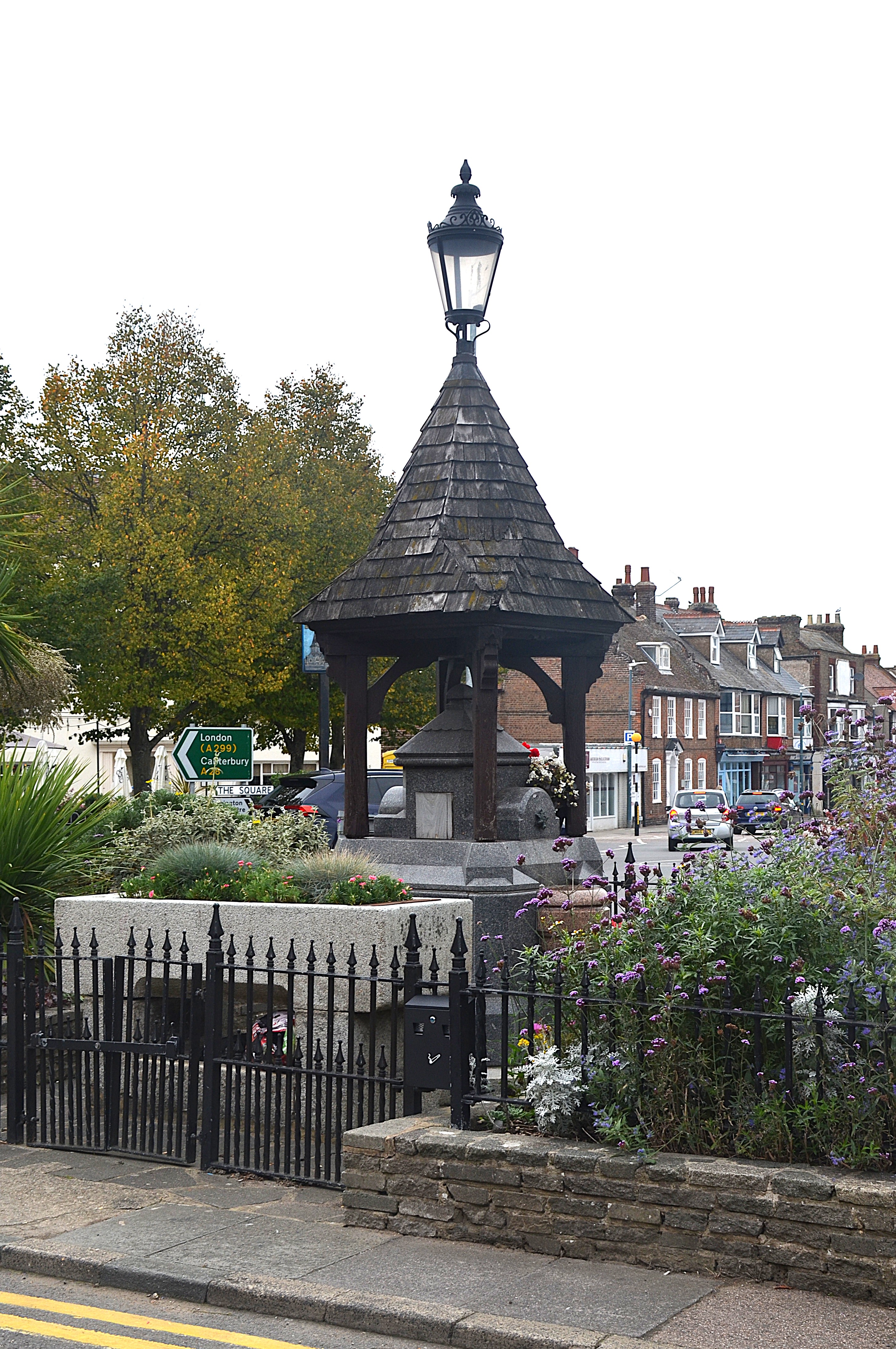 Fountain in centre with wooden canopy above topped by a lamp. Trees to left, buildings in background to right.