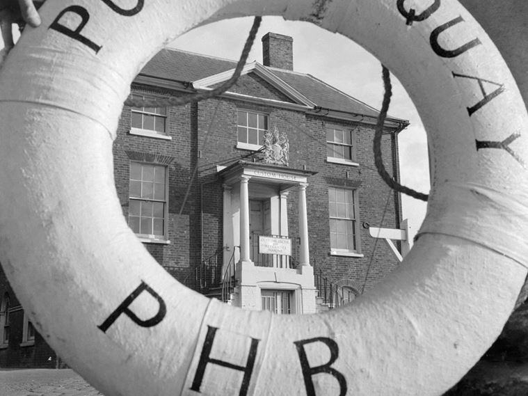 A view through a life ring, labelled "Poole Quay PHB", to the Custom House in Poole. The porch of the Custom's House has Tuscan columns, above which there is a cast-iron Royal Coat of Arms.