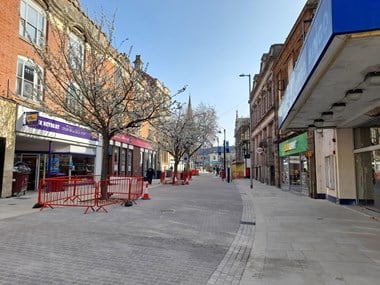 View along a pedestrianised high street with barriers and bollards in place around trees growing out of the paving.