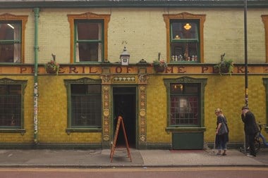 A colour photograph featuring part of a colourful public house frontage. The lower part of the building is covered in shiny tiles. Above is brickwork. Signage, partly hidden behind hanging baskets of flowers, reads: Peveril of the Peak'. Two people walk towards the entrance.