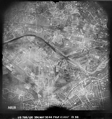 A black and white vertical aerial photograph of an urban area, featuring buildings, roads and a waterway that winds through the centre of the image. 