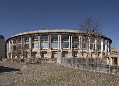 A view of the outside of a drum-shaped, two story building with columns supporting a roof structure and large windows.