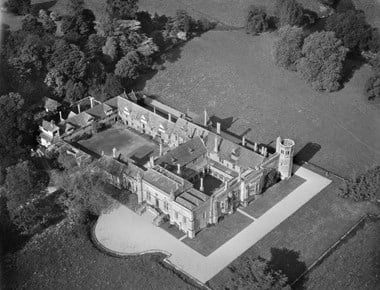 Black and white oblique aerial photograph of a multi-phase site set around a courtyard and a smaller cloister. Dominant in one corner is an octagonal tower that projects out beyond two elevations of the building. Neat paths and lawns front the two sides closest to the camera. Beyond a ha-ha are untended open areas and at the far side of the larger courtyard, to the top-left of the photograph, are clumps of trees.