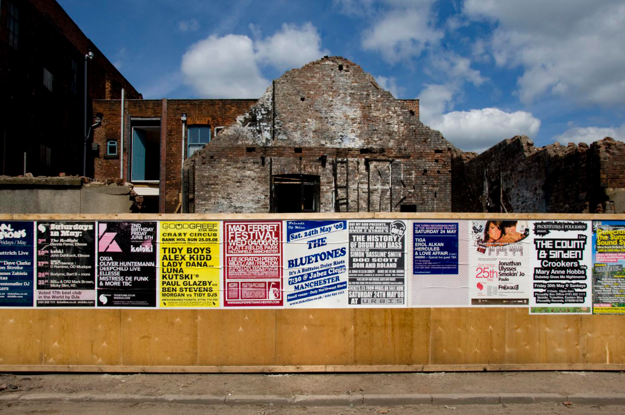 A wooden wall covered in colorful event posters with a partly demolished building and blue sky in the background.