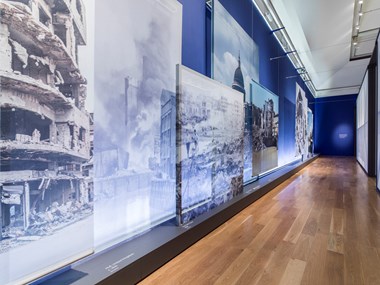 Large prints of wartime damage to buildings in England, hanging along a gallery wall and in places partially overlapping, as part of Historic England's What Remains exhibition at Imperial War Museum.