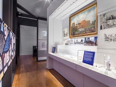 Photos, illustrations, a framed painting and artefacts displayed along a length of wall together with interpretation panels at the What Remains exhibition at the Imperial War Museum.