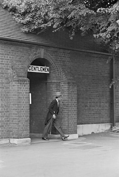 A man in top hat and tails strolling out of the arched entrance of the Gentlemen’s toilet, housed within a brick built structure.