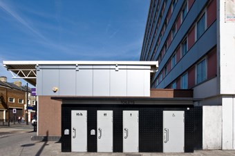 A row of four stainless steel doors, set within a black rectangular building, give access (left to right) to baby changing facilities; a unisex toilet a men’s toilet and a disabled toilet. The conveniences are surrounded by modern buildings in an urban setting.