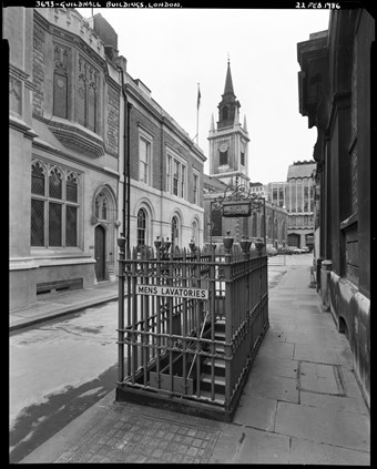 A view looking from the pavement beside the iron railings marking the entrance to the subterranean lavatories outside the Guildhall. A sign on the railings reads ‘MENS LAVATORIES’.  The Guildhall is to the right of the lavatory entrance and the Mayor’s and City of London Court is to the left.