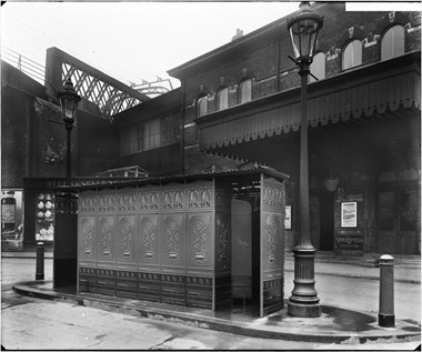 A black and white photo showing the exterior of a rectangular cast iron urinal building with gas lamp posts at either end. Brixton railway station in the background and a viaduct partially visible to the left.