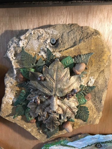 A stone roof tile or shingle decorated by a 21st-century artist in a painted relief motif depicting an oak leaf with  a human face, surrounded by acorns and foliage.