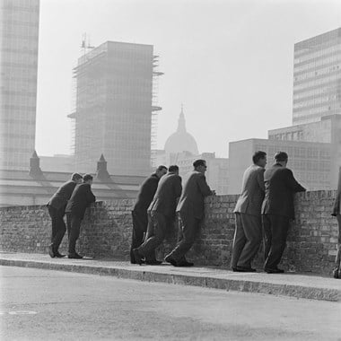 A black and white photo showing a group of men in suits leaning against a brick wall. The background shows the dome of St Paul’s Cathedral, as well as office blocks.