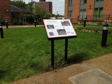 A photograph of a public information panel in its location in Stockport, situated on an area of grass surrounded buildings.