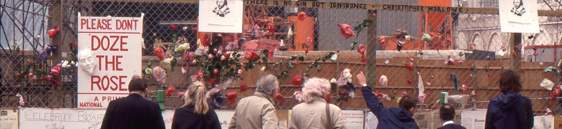 A photograph of seven members of the public looking at the excavation site of the Rose Theatre in London, behind a chain link fence.