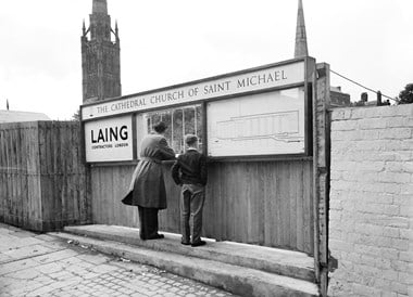 A man and boy looking through a viewing window at the construction site for Coventry Cathedral, with the Laing logo displayed on the left side, an elevation drawing of the cathedral displayed on the right side and a sign overhead displaying the cathedral name.