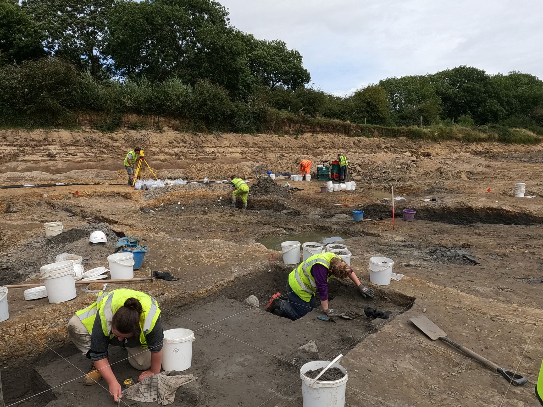 A group of archaeologists excavating: in the foreground two archaeologists are working within a 1 metre grid marked by strings.