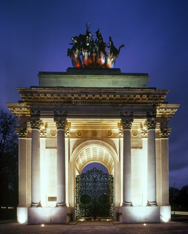 a floodlit arch with the night sky and trees behind it. There is a metal gate in the centre of the arch